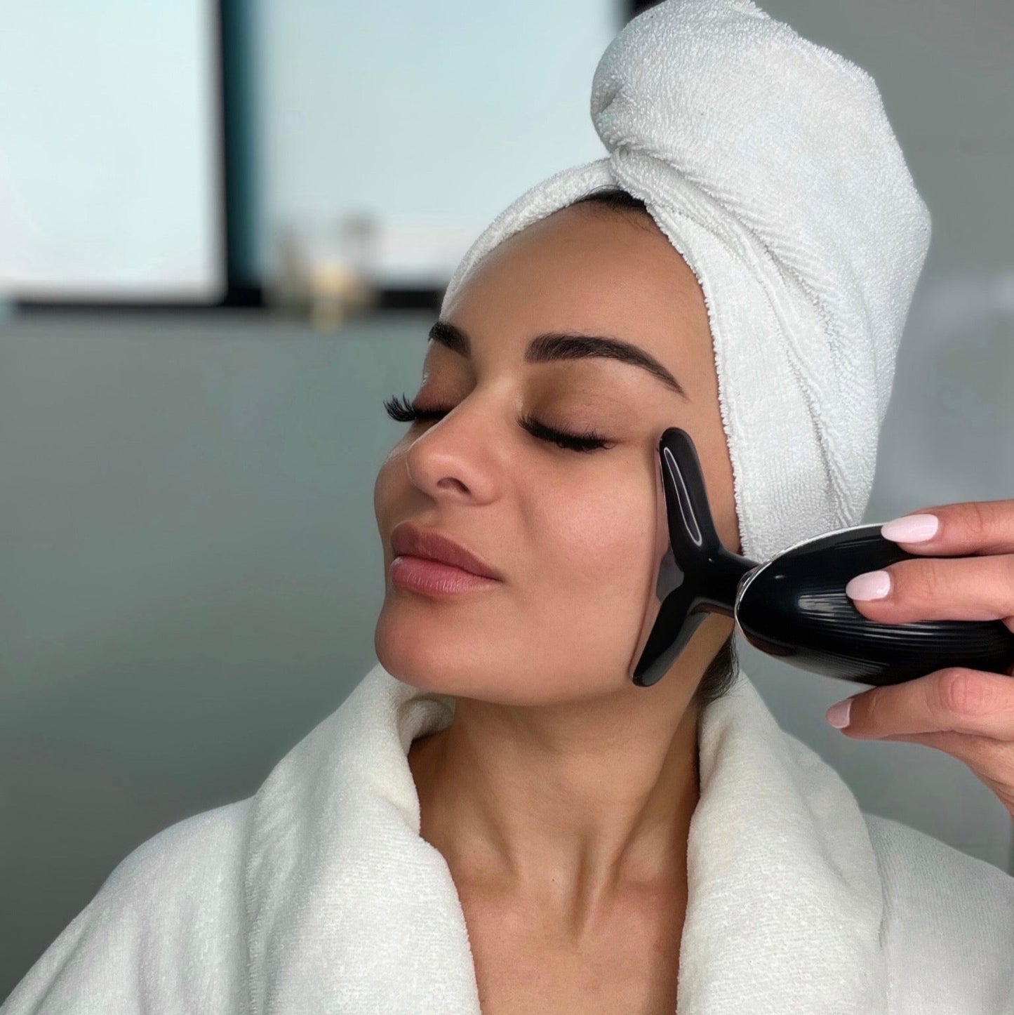 Woman in a white robe using a black handheld device on her face with a blurred indoor background