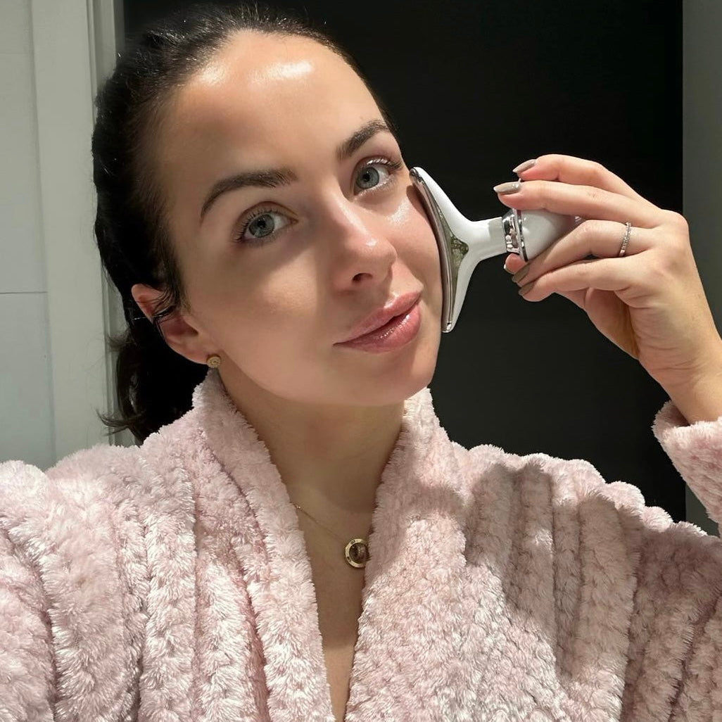 Woman in a pink robe using a facial roller device on her face.